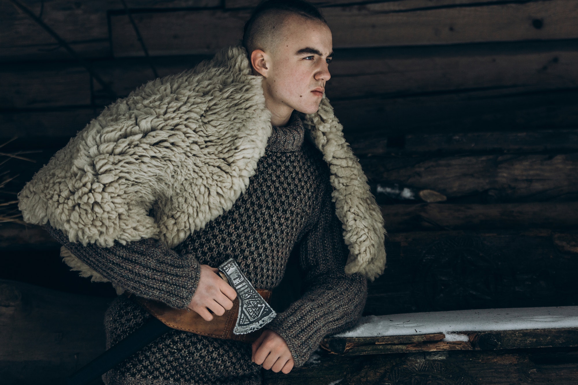 Ancient viking warrior with steel axe posing near wooden hut in scandinavian forest